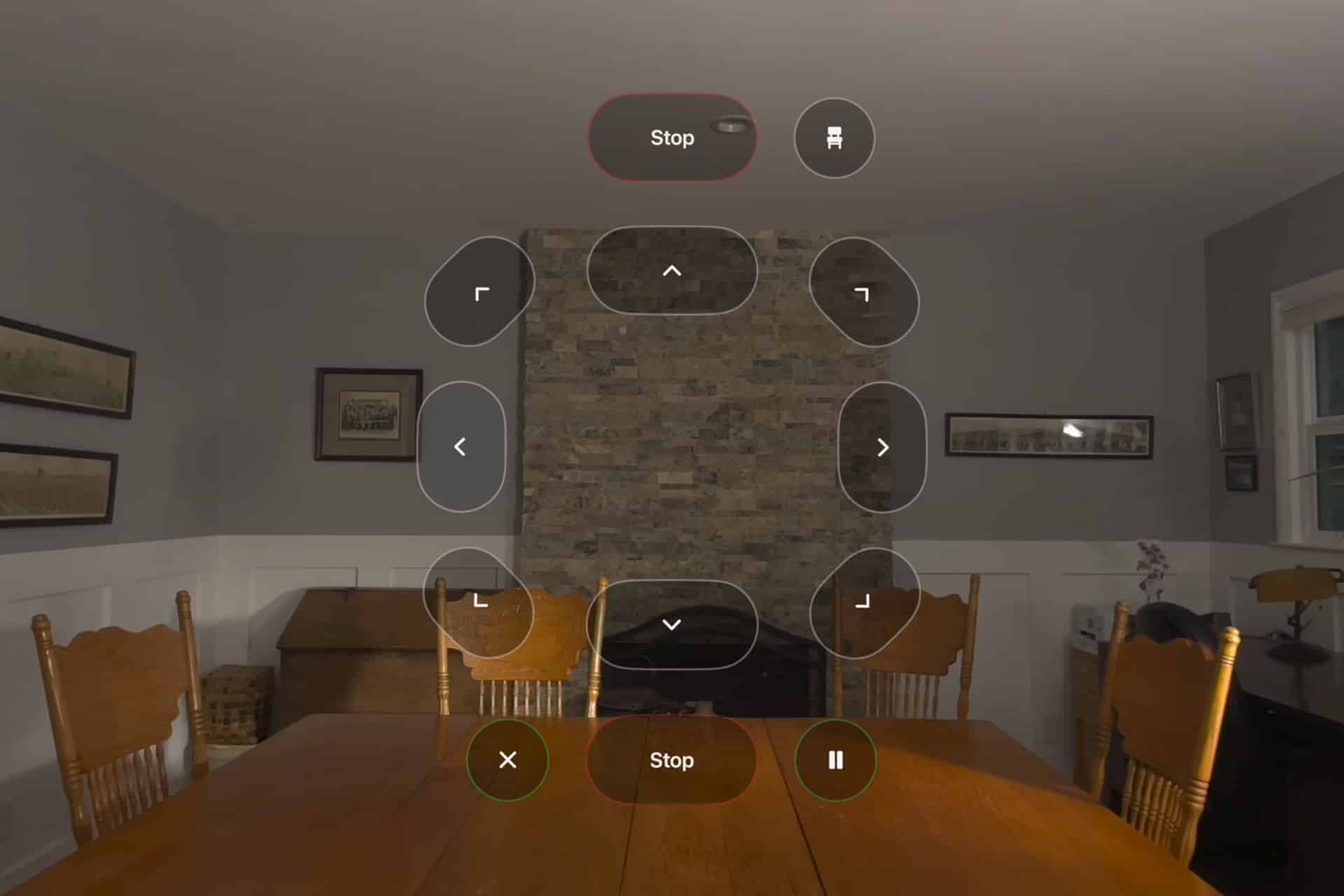 A dining room with wooden chairs and table, stone fireplace, framed pictures on gray walls, and an overlay of Vision Pro wheelchair control buttons displayed in the center of the image.