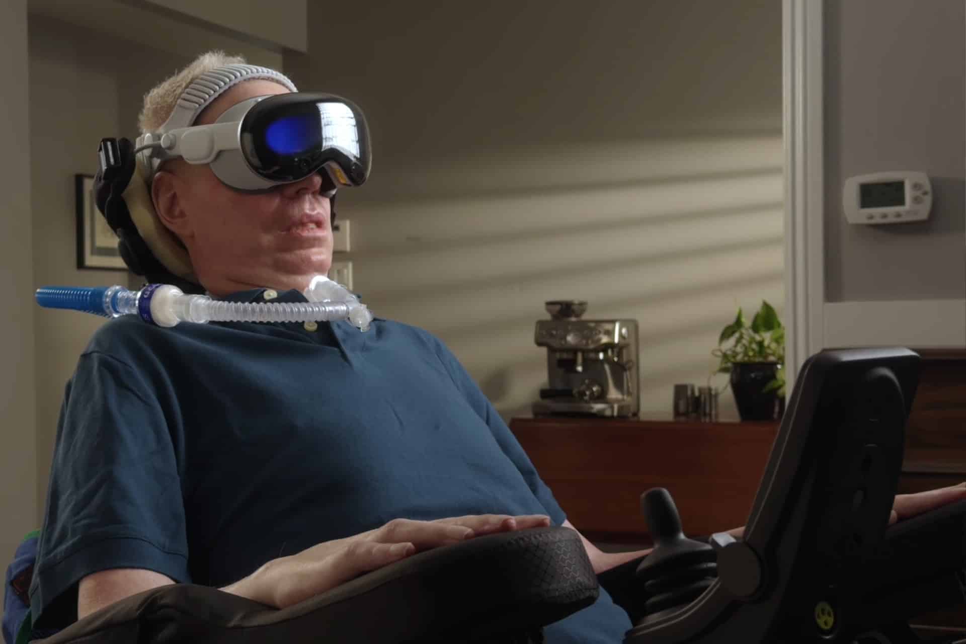 A person in a wheelchair wears a Vision Pro headset and uses a ventilator. They are indoors, with sunlight streaming through window blinds onto the wall behind them.