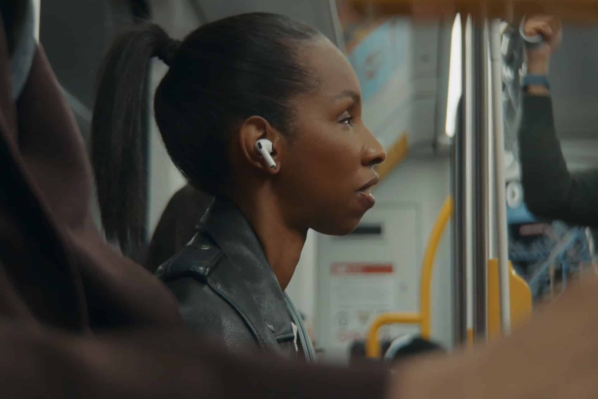 A woman with a ponytail, wearing a black leather jacket and wireless earbuds, sits on public transit using VoiceOver. The background shows part of the bus or train interior with yellow poles.