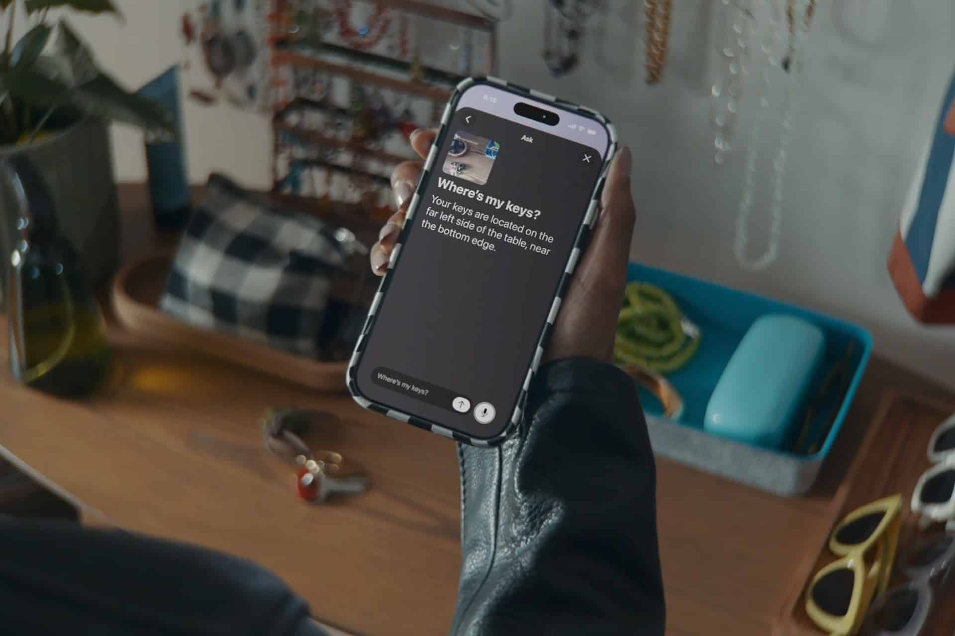A person holds a smartphone displaying a “Where’s my keys?” screen with instructions using VoiceOver. On the table in the background are keys, jewelry, a glasses case, and other personal items.
