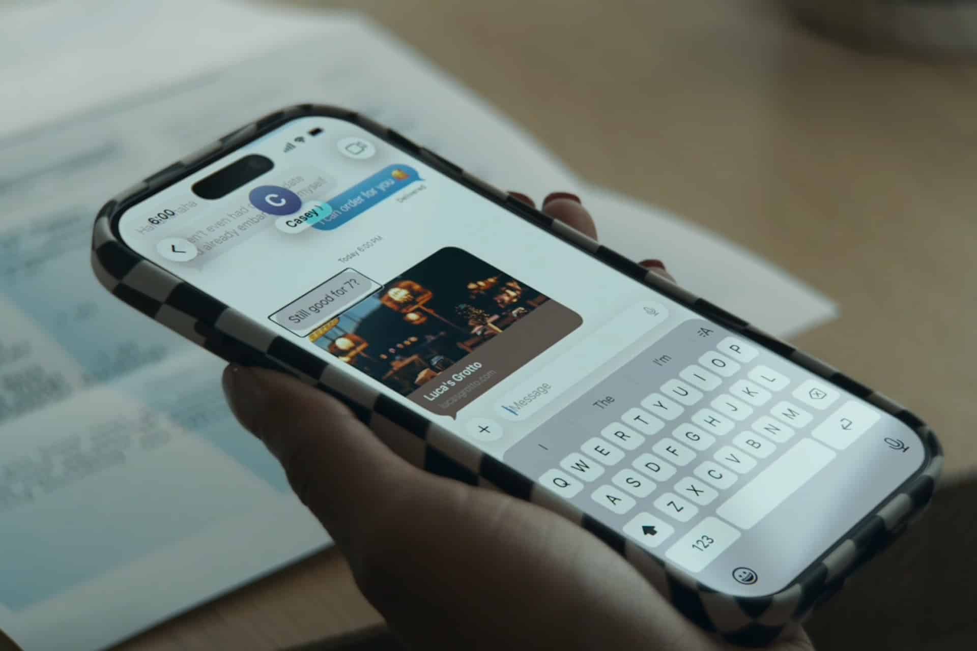 A person holds a smartphone with a checkered case, typing a message in a chat. The screen, accessed with VoiceOver, shows a GIF and the text “Sit pack by 2!!!” while documents are blurred in the background.