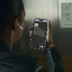 A person wearing a black jacket and wireless earbud holds up a smartphone, using an accessibility app to read a handwritten "OUT OF ORDER USE SOUTH ELEVATOR" sign taped to an elevator door.