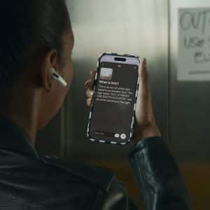 A person wearing a black jacket and wireless earbud holds up a smartphone, using an accessibility app to read a handwritten "OUT OF ORDER USE SOUTH ELEVATOR" sign taped to an elevator door.