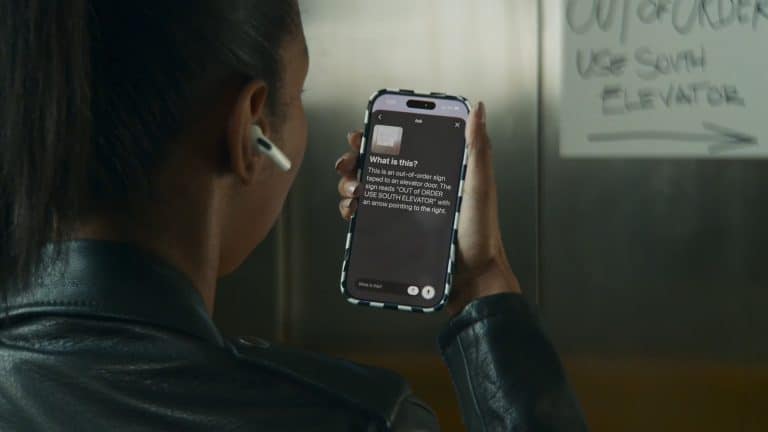 A person wearing a black jacket and wireless earbud holds up a smartphone, using an accessibility app to read a handwritten "OUT OF ORDER USE SOUTH ELEVATOR" sign taped to an elevator door.