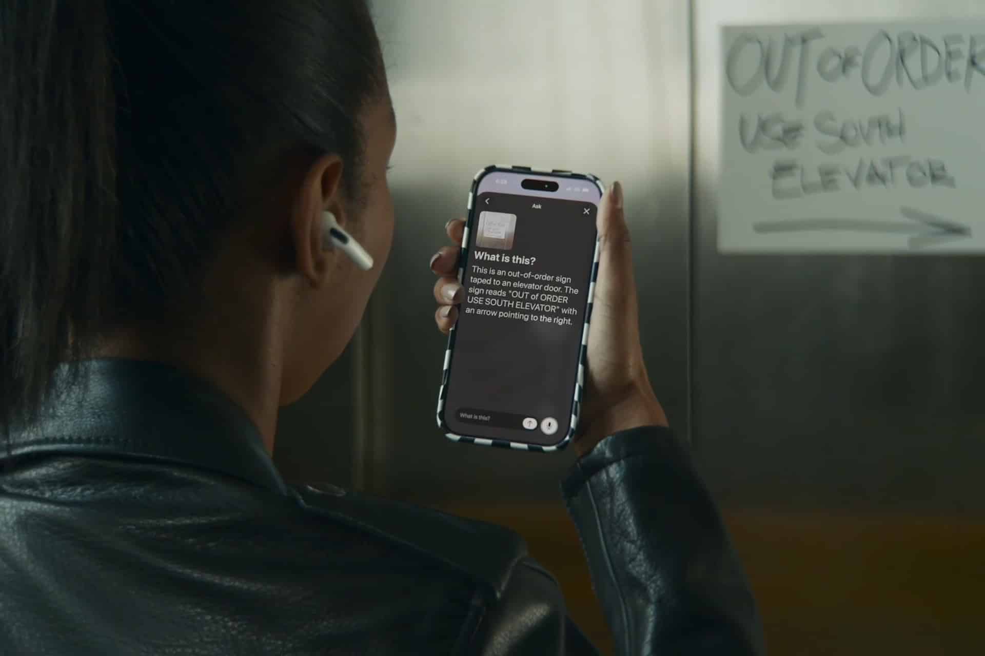 A person wearing a black jacket and wireless earbud holds up a smartphone, using an accessibility app to read a handwritten "OUT OF ORDER USE SOUTH ELEVATOR" sign taped to an elevator door.