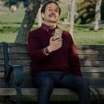 A man wearing AirPods sits on a park bench holding an iPhone, with a dog partially visible beside him.