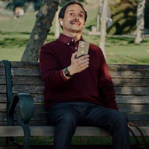 A man wearing AirPods sits on a park bench holding an iPhone, with a dog partially visible beside him.