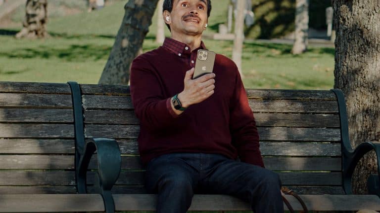 A man wearing AirPods sits on a park bench holding an iPhone, with a dog partially visible beside him.