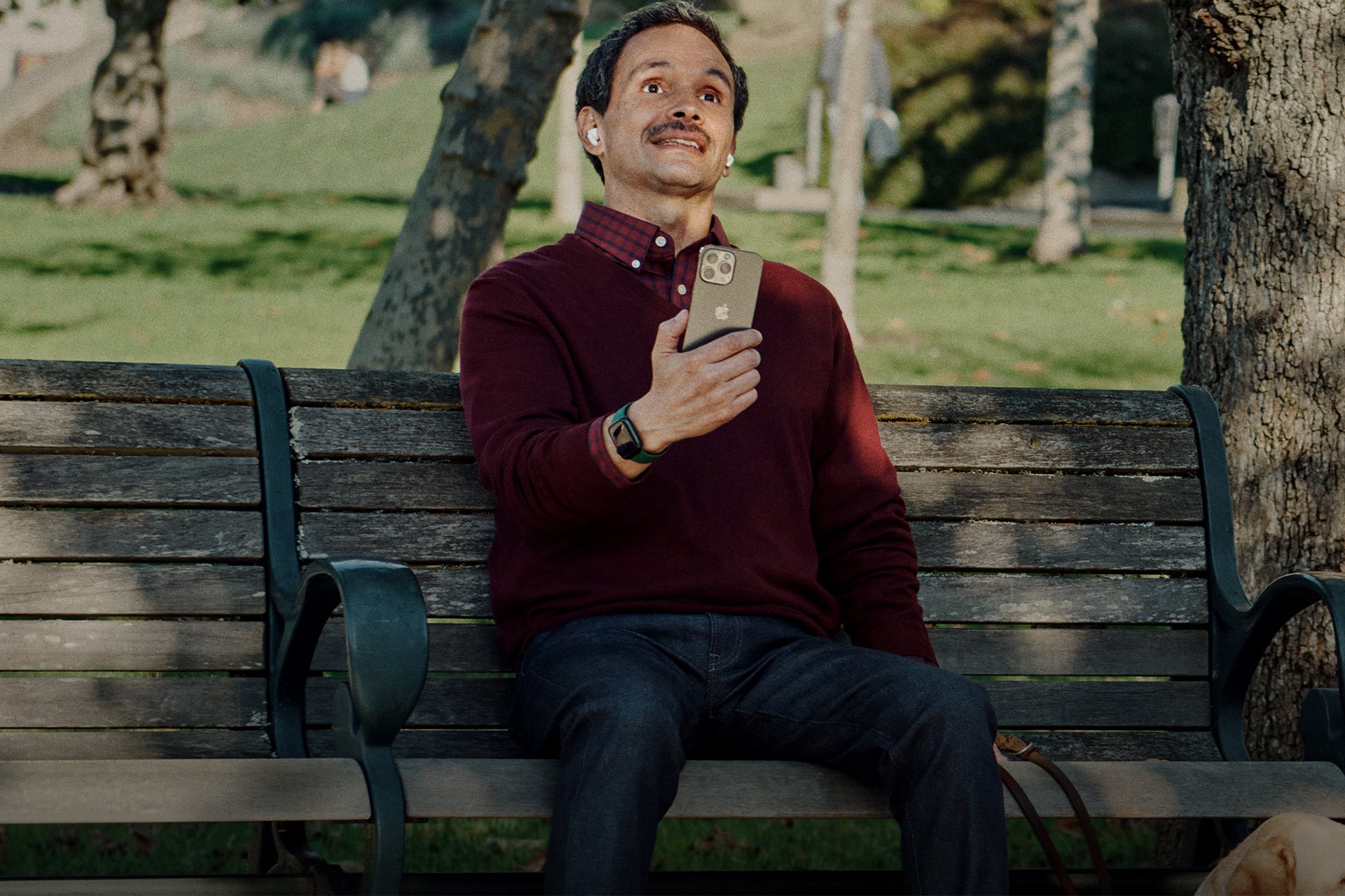 A man wearing AirPods sits on a park bench holding an iPhone, with a dog partially visible beside him.