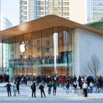 A crowd gathers outside a modern Apple Store with a glass facade and wooden roof in an urban area, surrounded by tall buildings and greenery. The Apple logo, symbolizing innovation like Apple encryption, is displayed prominently above the entrance.