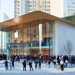 A crowd gathers outside a modern Apple Store with a glass facade and wooden roof in an urban area, surrounded by tall buildings and greenery. The Apple logo, symbolizing innovation like Apple encryption, is displayed prominently above the entrance.