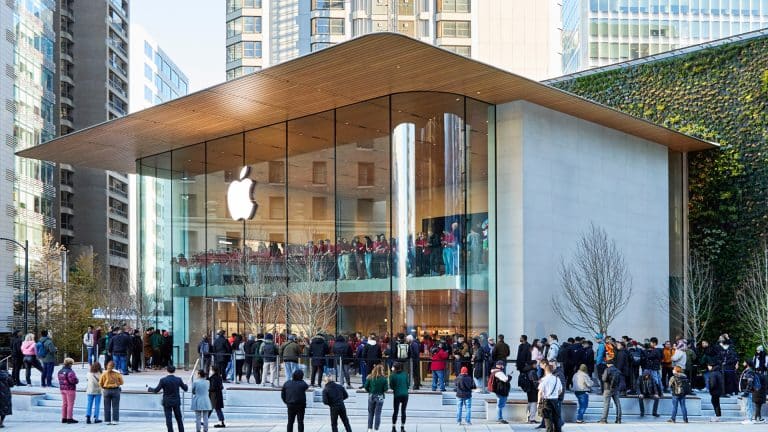 A crowd gathers outside a modern Apple Store with a glass facade and wooden roof in an urban area, surrounded by tall buildings and greenery. The Apple logo, symbolizing innovation like Apple encryption, is displayed prominently above the entrance.