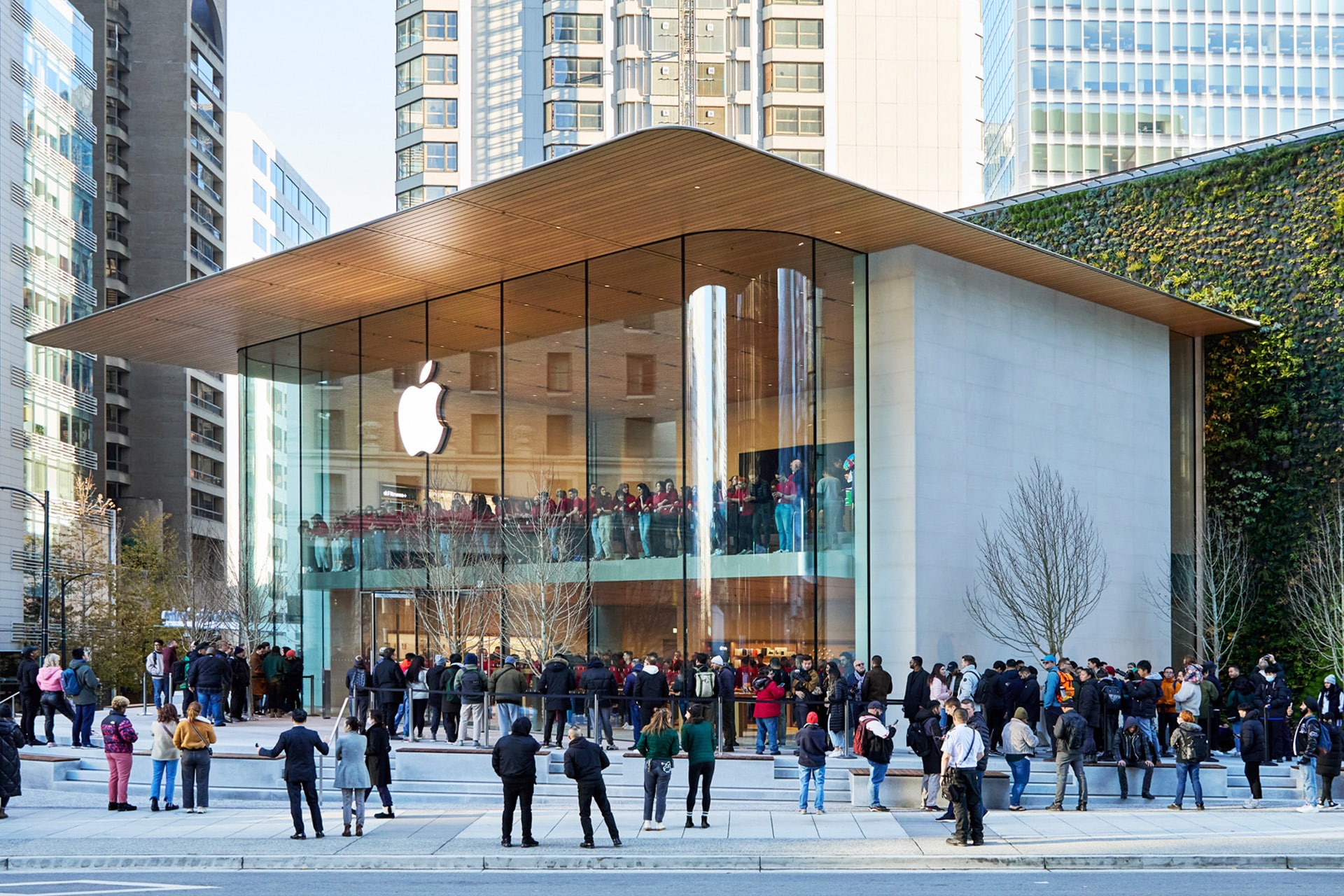 A crowd gathers outside a modern Apple Store with a glass facade and wooden roof in an urban area, surrounded by tall buildings and greenery. The Apple logo, symbolizing innovation like Apple encryption, is displayed prominently above the entrance.