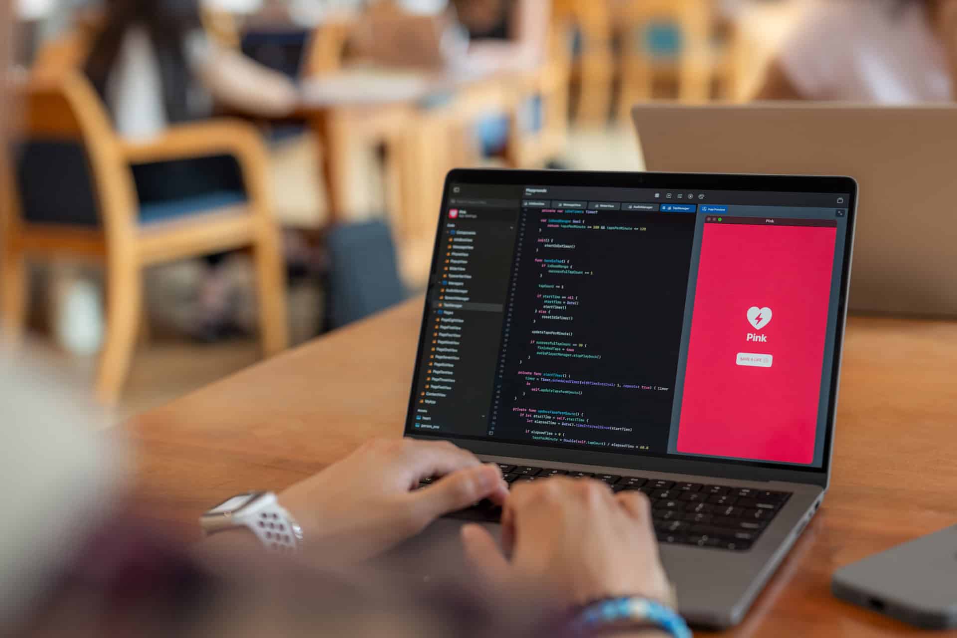 A person is working on a laptop displaying code in Swift Playgrounds on one side and a mobile app interface with a heart icon and the word "Pink" on a pink background on the other, set in a busy workspace.