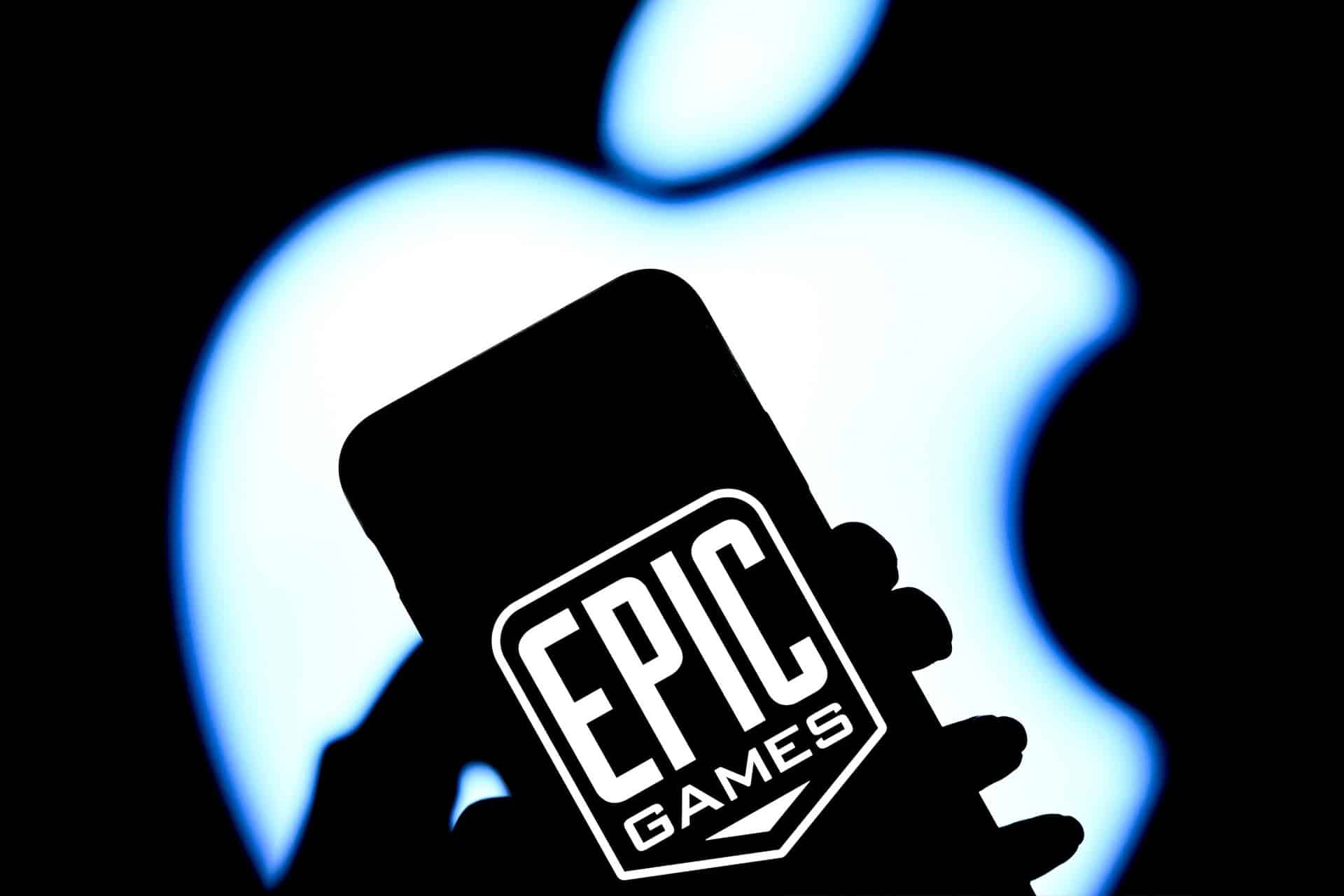 A hand holds a phone displaying the Epic Games logo in white, with a large, blurred Apple logo illuminated in the background—hinting at their ongoing battle over App Store fees.