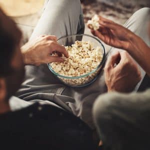 Two people sitting close together, sharing a glass bowl of popcorn while enjoying a movie with Apple TV Dolby Vision. Only their arms and hands are clearly visible as they reach for the popcorn, suggesting a relaxed, casual atmosphere.