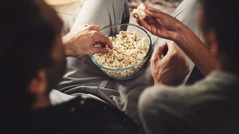 Two people sitting close together, sharing a glass bowl of popcorn while enjoying a movie with Apple TV Dolby Vision. Only their arms and hands are clearly visible as they reach for the popcorn, suggesting a relaxed, casual atmosphere.
