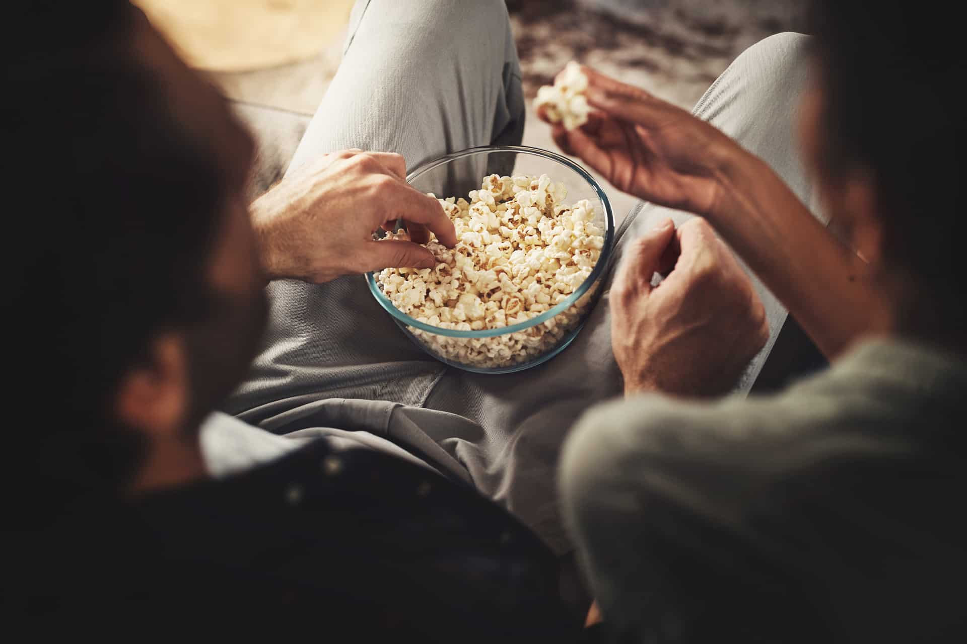 Two people sitting close together, sharing a glass bowl of popcorn while enjoying a movie with Apple TV Dolby Vision. Only their arms and hands are clearly visible as they reach for the popcorn, suggesting a relaxed, casual atmosphere.