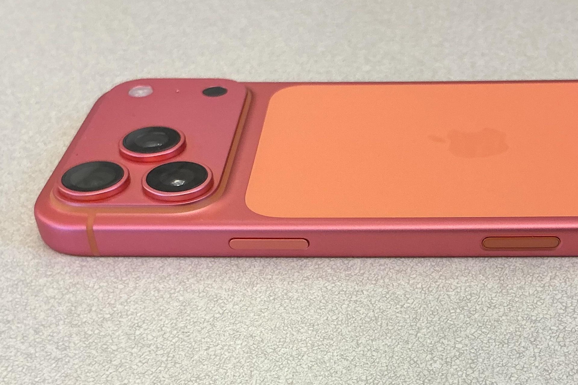 A close-up of the back and side of an orange turning pink iPhone 17 Pro placed on a light gray surface, highlighting its dual camera lenses and side buttons.