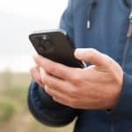 A person wearing a blue jacket is holding a black smartphone with both hands outdoors, possibly checking satellite coverage amid greenery and sky—ideal for staying connected in iPhone dead zones.