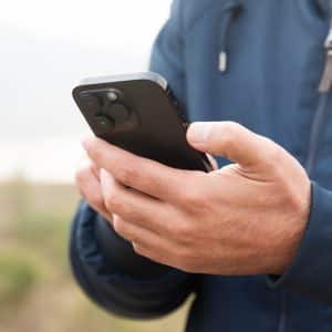 A person wearing a blue jacket is holding a black smartphone with both hands outdoors, possibly checking satellite coverage amid greenery and sky—ideal for staying connected in iPhone dead zones.