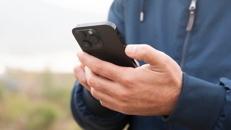 A person wearing a blue jacket is holding a black smartphone with both hands outdoors, possibly checking satellite coverage amid greenery and sky—ideal for staying connected in iPhone dead zones.