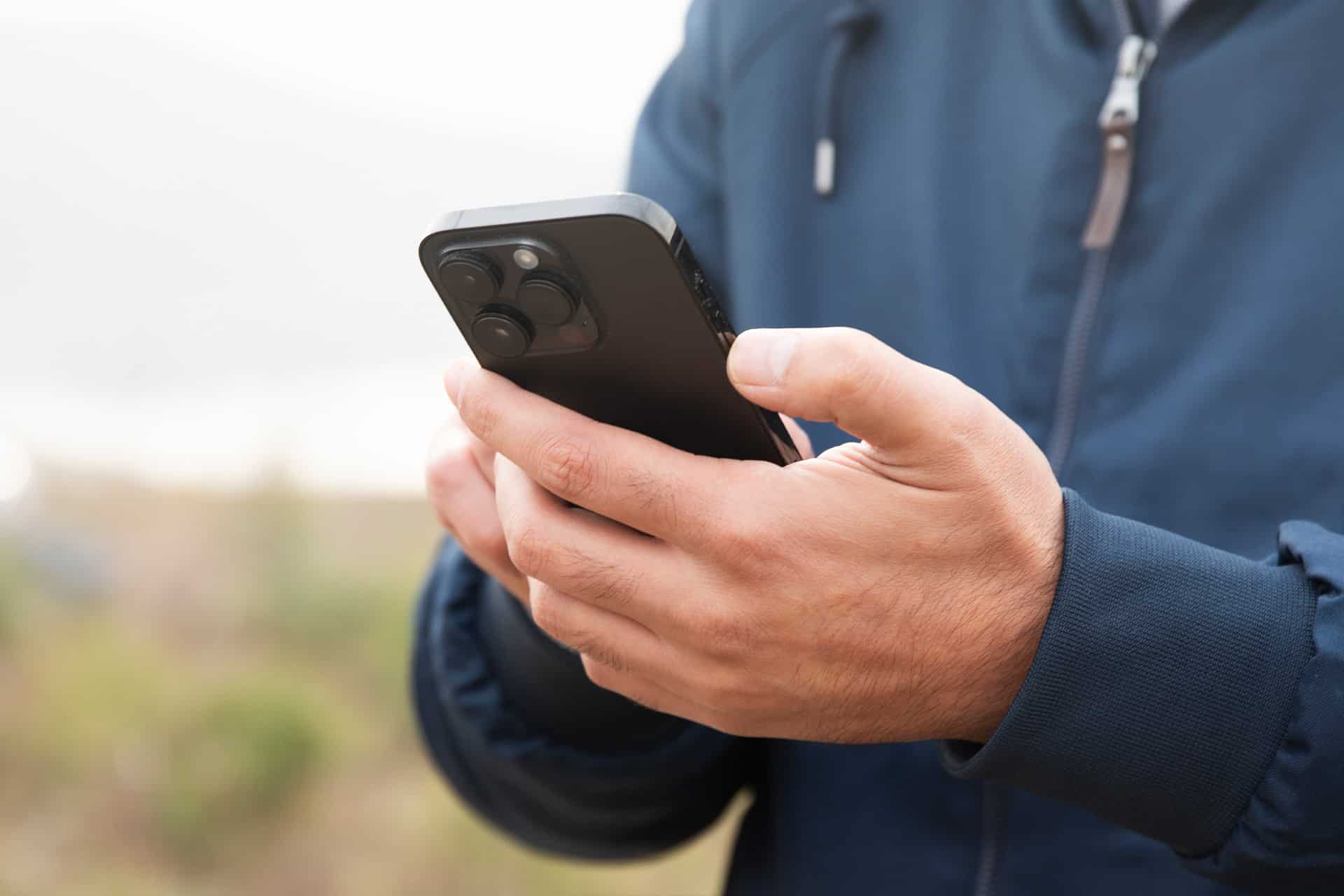 A person wearing a blue jacket is holding a black smartphone with both hands outdoors, possibly checking satellite coverage amid greenery and sky—ideal for staying connected in iPhone dead zones.
