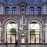 A large, ornate stone building with tall arched windows and a central entrance displays a white flag in the UK. People walk past, while inside are wooden tables, benches, and green trees—a calm scene amidst news of the Apple iCloud lawsuit.