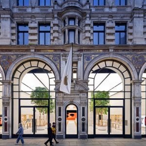 A large, ornate stone building with tall arched windows and a central entrance displays a white flag in the UK. People walk past, while inside are wooden tables, benches, and green trees—a calm scene amidst news of the Apple iCloud lawsuit.