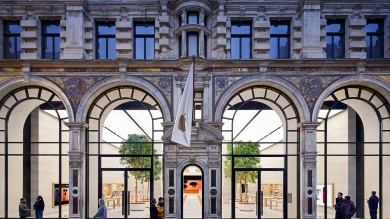A large, ornate stone building with tall arched windows and a central entrance displays a white flag in the UK. People walk past, while inside are wooden tables, benches, and green trees—a calm scene amidst news of the Apple iCloud lawsuit.