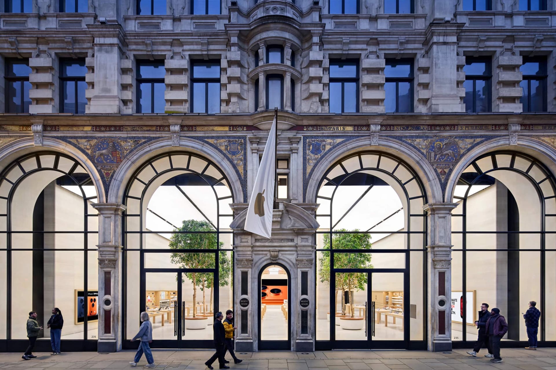A large, ornate stone building with tall arched windows and a central entrance displays a white flag in the UK. People walk past, while inside are wooden tables, benches, and green trees—a calm scene amidst news of the Apple iCloud lawsuit.