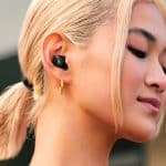 A person with blond hair in a low ponytail is wearing a black Beats Studio Buds wireless earbud and a gold hoop earring, looking content with eyes closed.