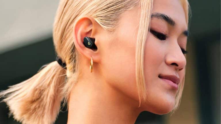 A person with blond hair in a low ponytail is wearing a black Beats Studio Buds wireless earbud and a gold hoop earring, looking content with eyes closed.