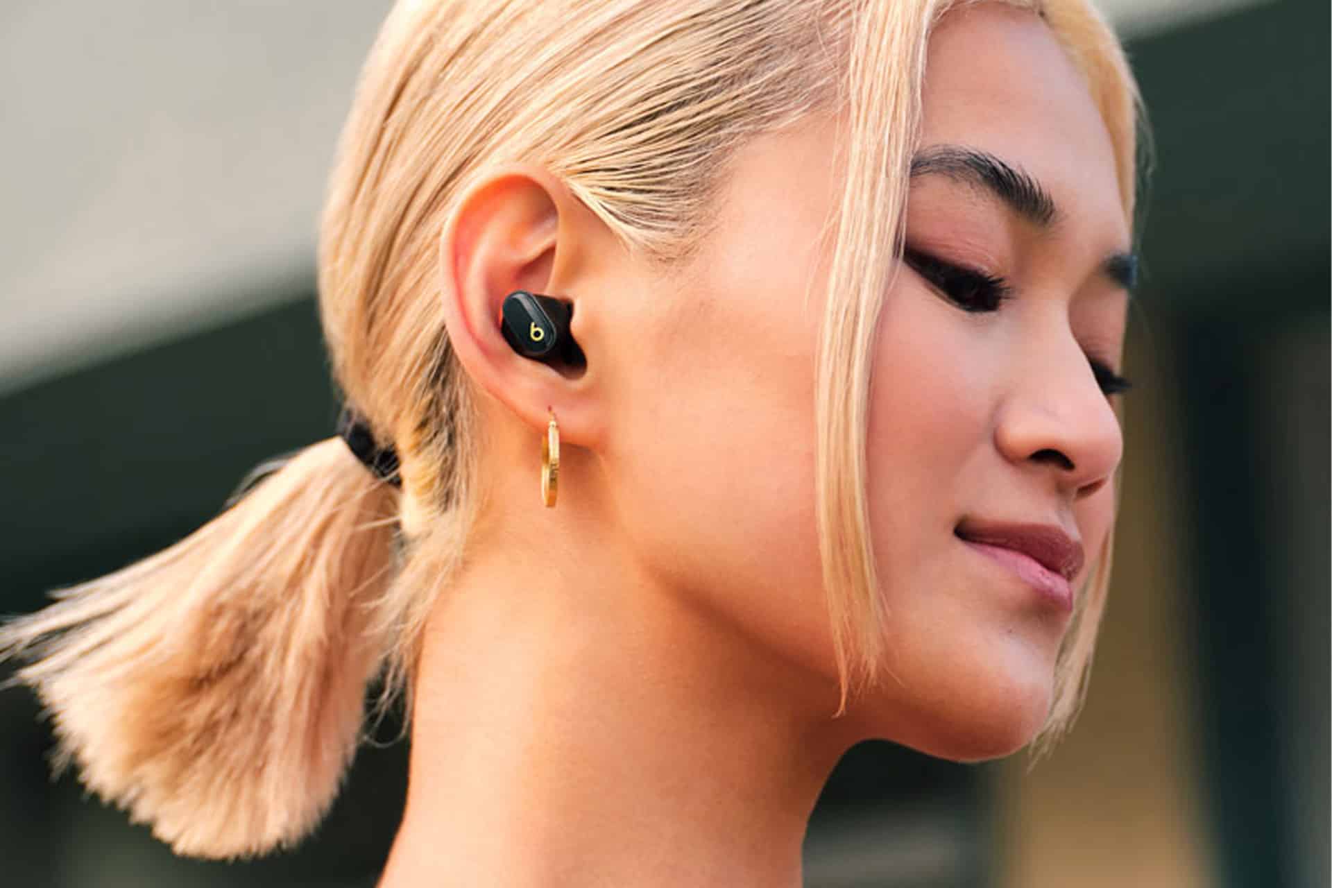 A person with blond hair in a low ponytail is wearing a black Beats Studio Buds wireless earbud and a gold hoop earring, looking content with eyes closed.