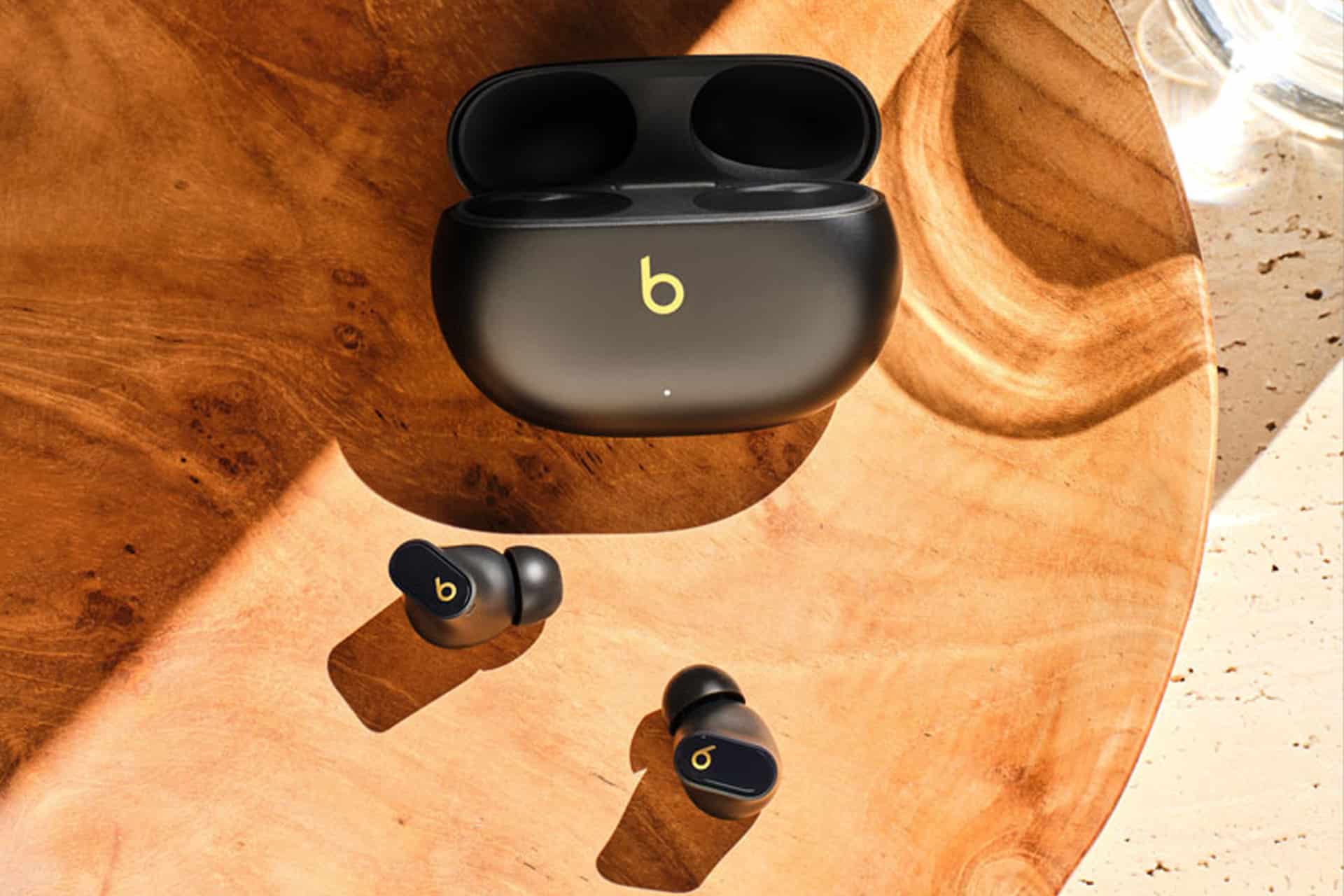 A pair of black Bluetooth wireless earbuds with gold “b” logos rests on a wooden surface beside an open charging case, also black with a gold “b” logo. Sunlight casts soft shadows across the scene.