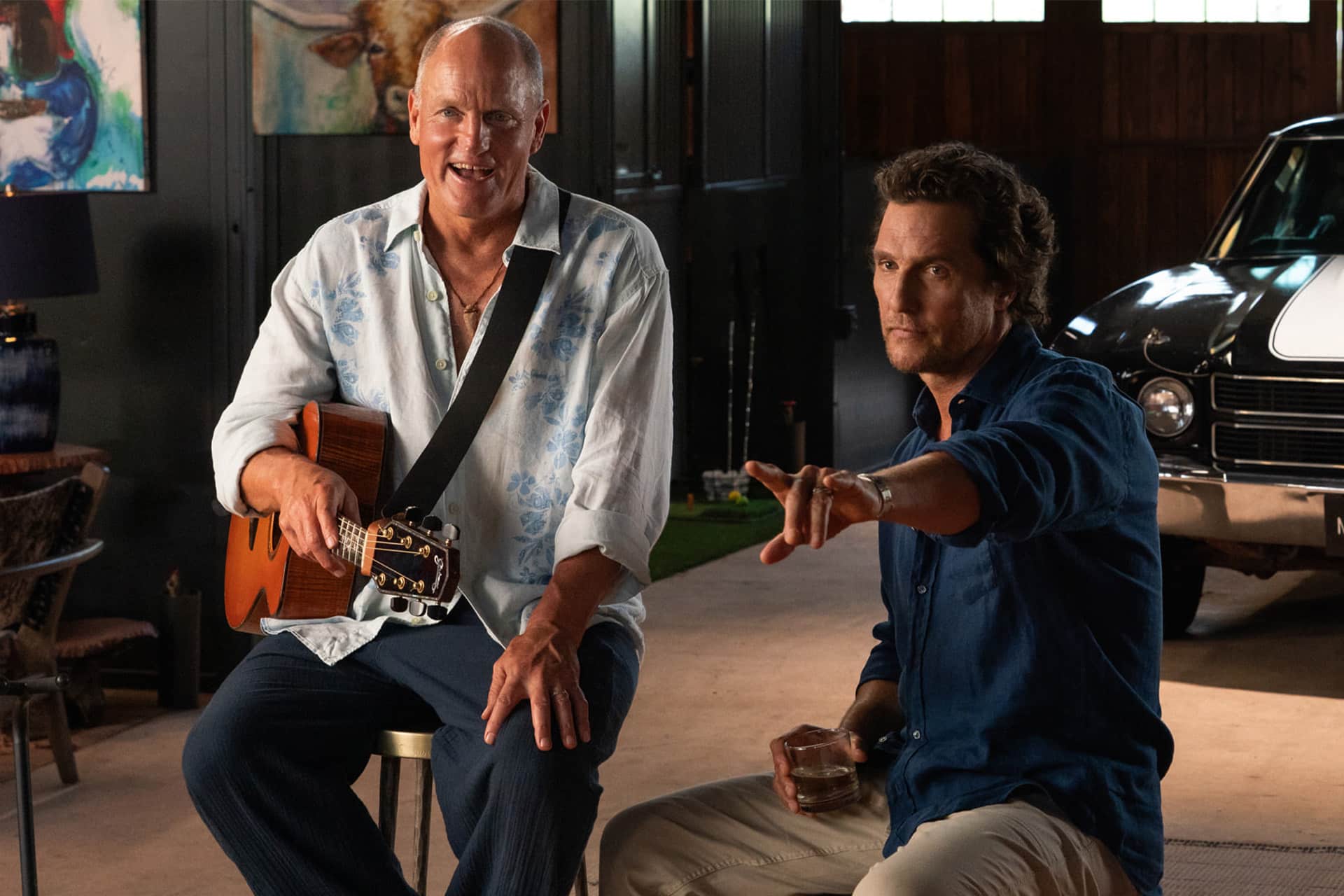 Brothers - Two brothers sit indoors; one, resembling Matthew McConaughey in a light floral shirt, smiles with a guitar, while the other gestures with his drink. A classic car peeks from the background—an Apple Originals vibe.
