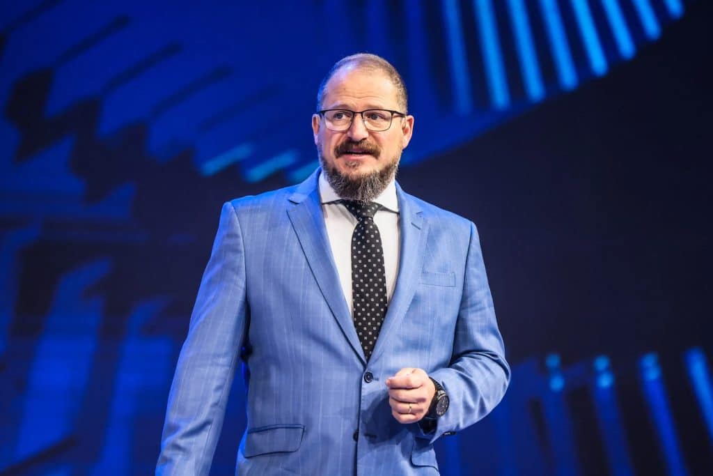 Cristiano Amon stands on stage in a blue suit, white shirt, and black polka dot tie, gesturing as he discusses the future of agentic AI and Qualcomm AI agents against a striking blue background. Glasses and a beard complete his confident look.