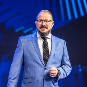 Cristiano Amon stands on stage in a blue suit, white shirt, and black polka dot tie, gesturing as he discusses the future of agentic AI and Qualcomm AI agents against a striking blue background. Glasses and a beard complete his confident look.