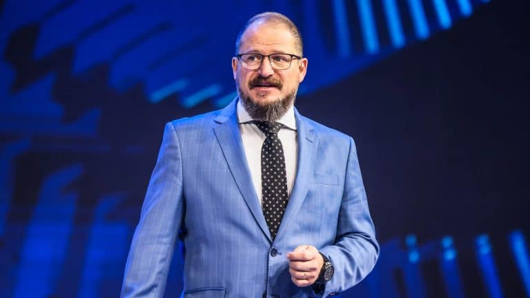 Cristiano Amon stands on stage in a blue suit, white shirt, and black polka dot tie, gesturing as he discusses the future of agentic AI and Qualcomm AI agents against a striking blue background. Glasses and a beard complete his confident look.