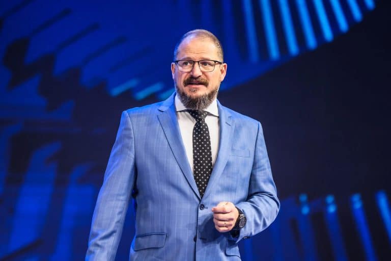 Cristiano Amon stands on stage in a blue suit, white shirt, and black polka dot tie, gesturing as he discusses the future of agentic AI and Qualcomm AI agents against a striking blue background. Glasses and a beard complete his confident look.