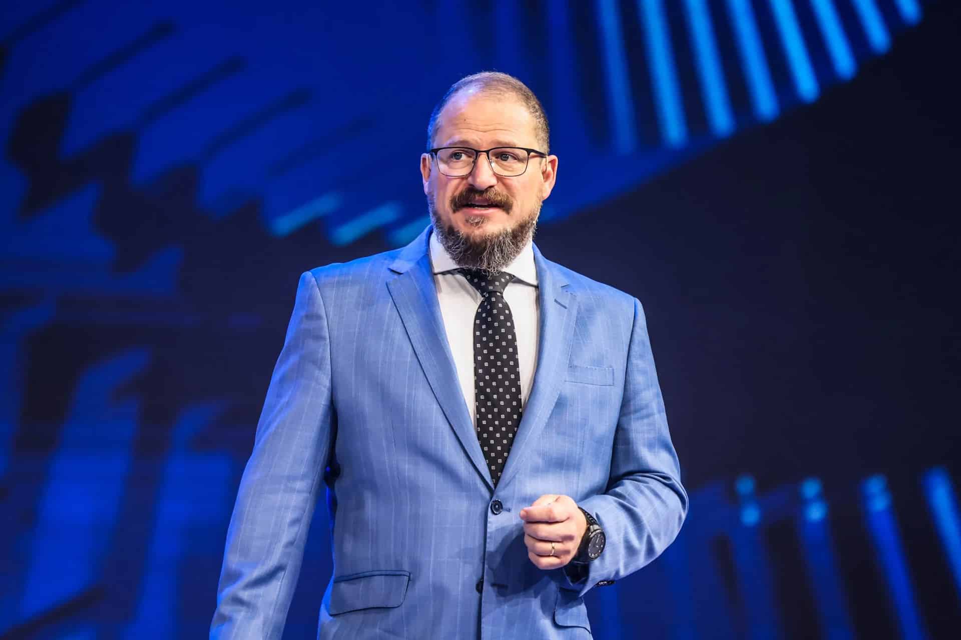 Cristiano Amon stands on stage in a blue suit, white shirt, and black polka dot tie, gesturing as he discusses the future of agentic AI and Qualcomm AI agents against a striking blue background. Glasses and a beard complete his confident look.