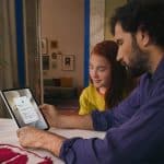 A man and a girl sit at a table looking at a tablet together. The screen displays an Apple parental controls login page. They are indoors with fruit and a plant on the table, creating a focused and collaborative atmosphere.
