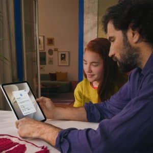 A man and a girl sit at a table looking at a tablet together. The screen displays an Apple parental controls login page. They are indoors with fruit and a plant on the table, creating a focused and collaborative atmosphere.