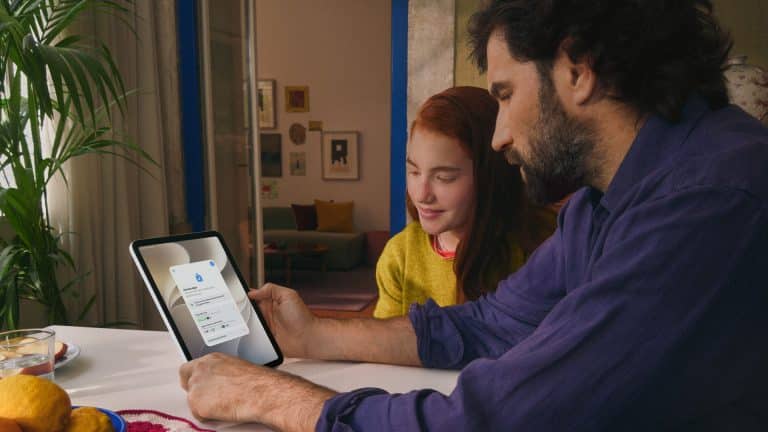 A man and a girl sit at a table looking at a tablet together. The screen displays an Apple parental controls login page. They are indoors with fruit and a plant on the table, creating a focused and collaborative atmosphere.