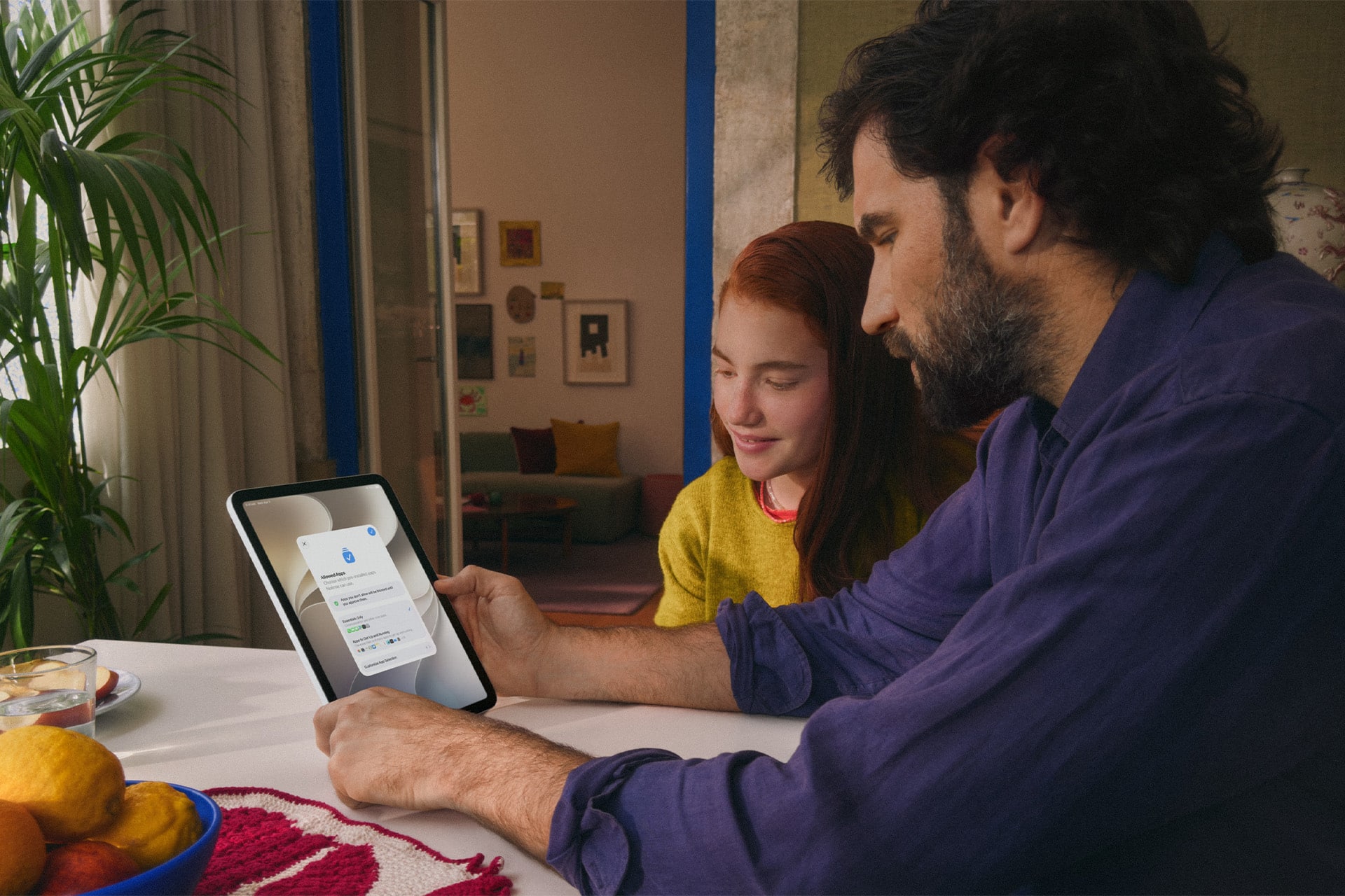 A man and a girl sit at a table looking at a tablet together. The screen displays an Apple parental controls login page. They are indoors with fruit and a plant on the table, creating a focused and collaborative atmosphere.