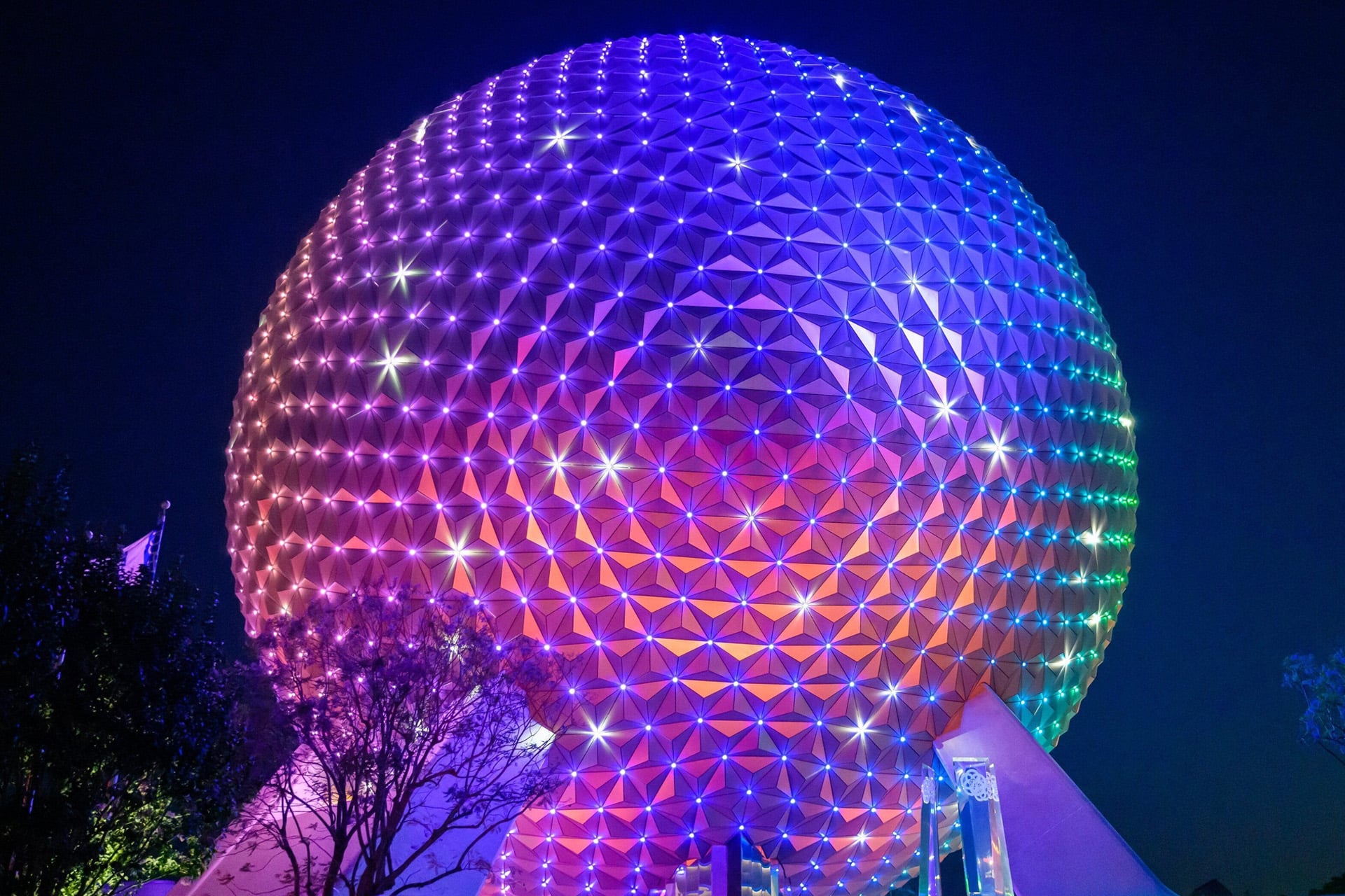 A large geodesic sphere at night, illuminated with vibrant, colorful lights in shades of purple, pink, blue, and green—reminiscent of a scene you’d catch on Apple TV. Trees are visible in front of the structure.
