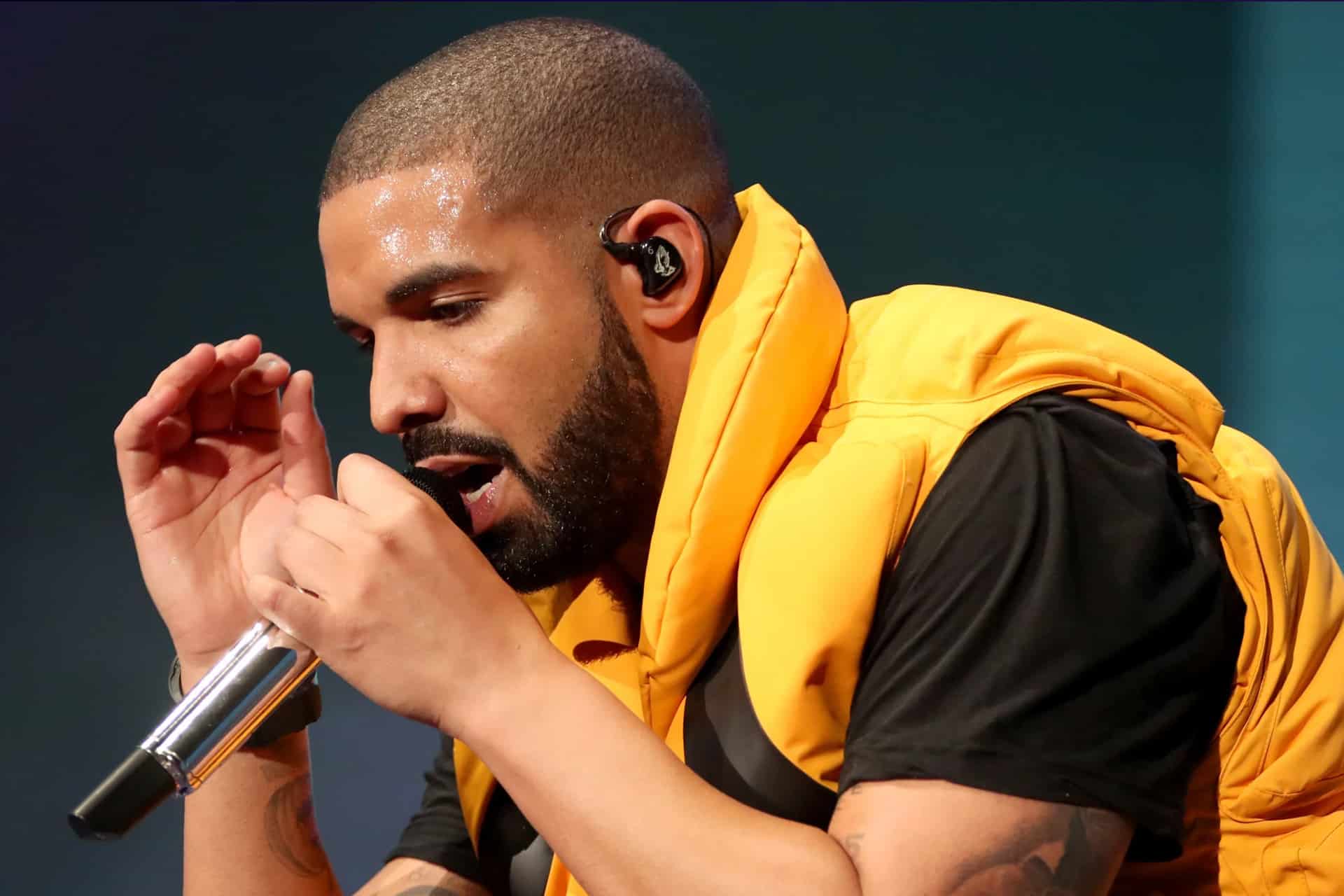 A man with a beard in a yellow vest holds a microphone and performs on stage, appearing focused and expressive, reminiscent of Drake during an Apple Music live session.