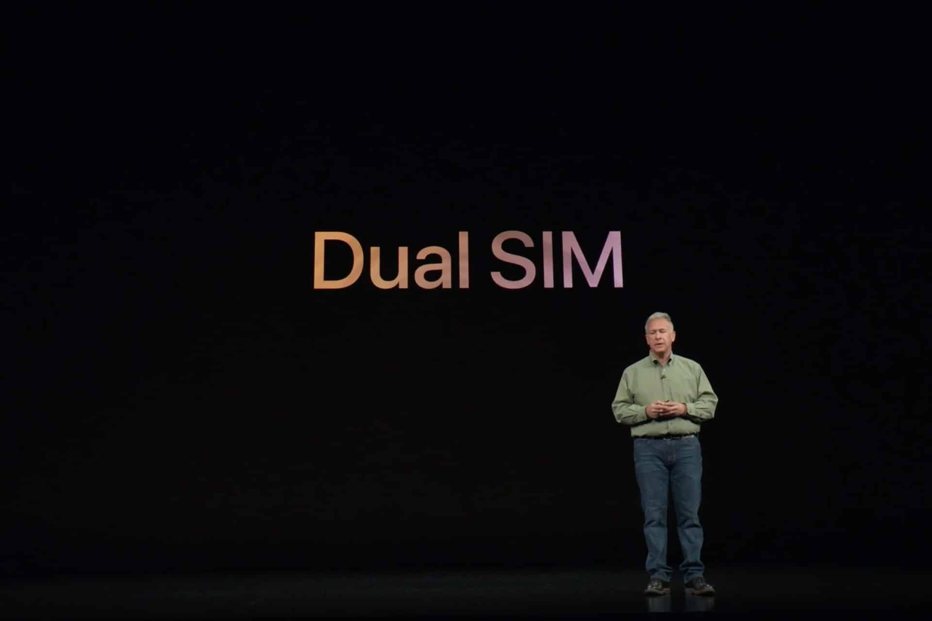 Dual SIM - A man in a green shirt and jeans stands on a dark stage beneath large text that reads "Dual SIM" in gradient colors, highlighting the convenience of keeping a personal line with iPhone eSIM support.