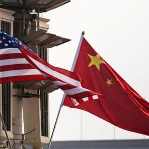 The American flag and the Chinese flag wave side by side in front of a beige guard tower or surveillance structure, highlighting tensions over memory chips and China memory firms against a pale sky.