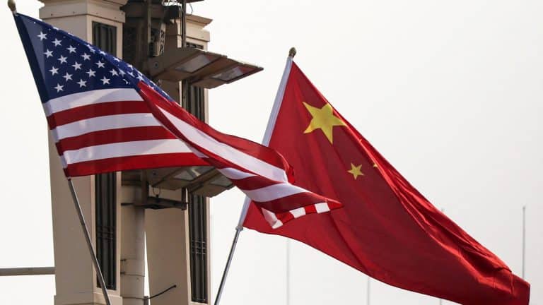 The American flag and the Chinese flag wave side by side in front of a beige guard tower or surveillance structure, highlighting tensions over memory chips and China memory firms against a pale sky.